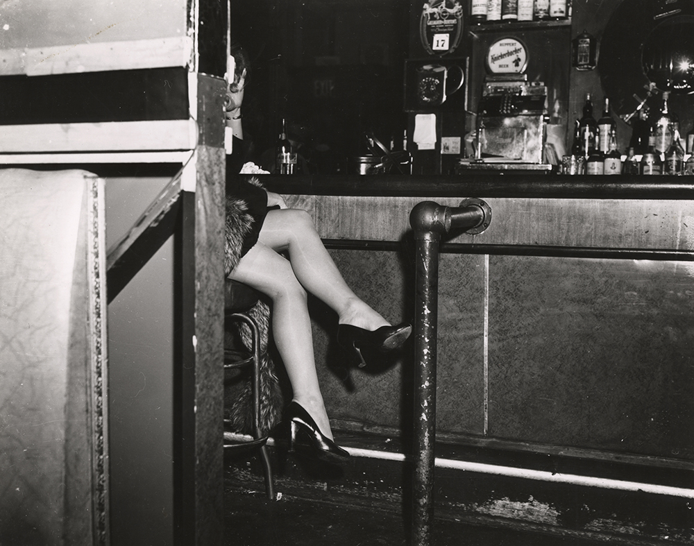 A woman is partially visible as she sits on a bar stool at a bar, her legs are crossed; in the background are bottles of liquor, New York, ca.1940s. (Photo by Weegee (Arthur Fellig)/International Center of Photography/Getty Images)