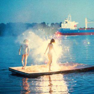 RYAN MCGINLEY, naked woman and man standing in sparklers on the water with a boat in the distance