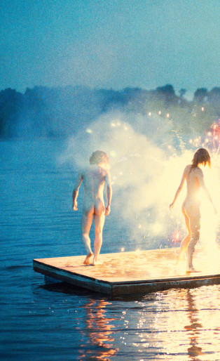 RYAN MCGINLEY, naked woman and man standing in sparklers on the water with a boat in the distance