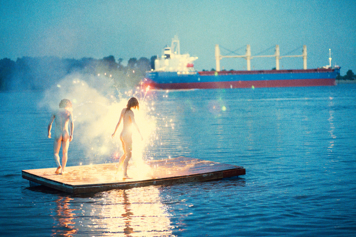 RYAN MCGINLEY, naked woman and man standing in sparklers on the water with a boat in the distance