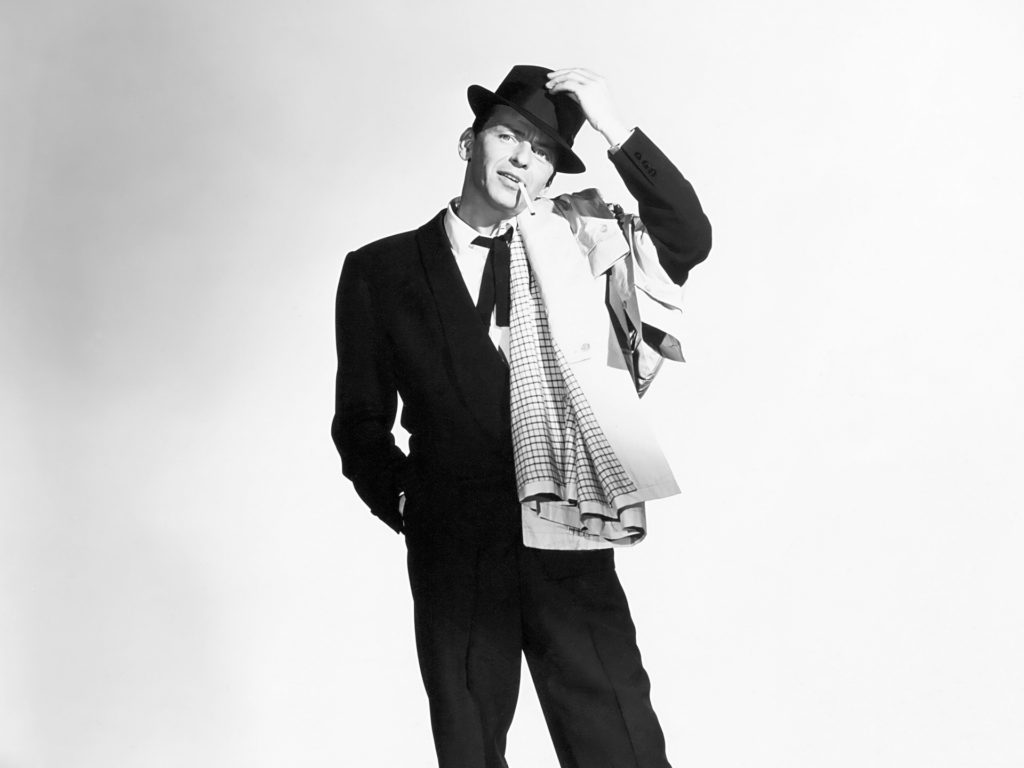 Classic photograph of Frank Sinatra wearing a black suit, hat and beige trench coat smoking a cigarette