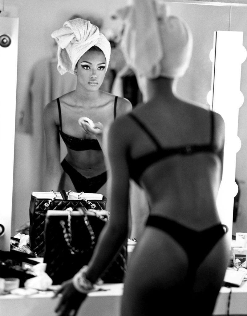 Black and white photograph of Naomi Campbell putting her make-up on wearing black bra and thong