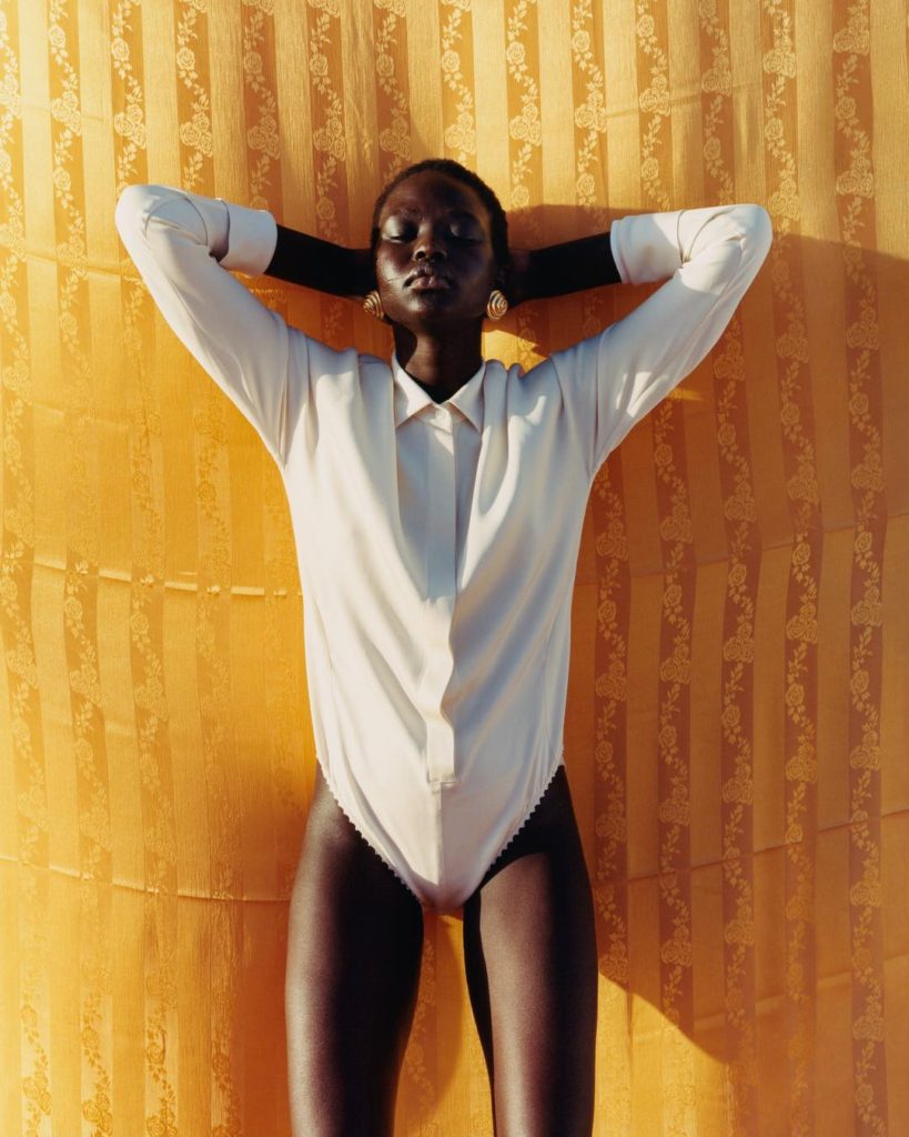 Tyler Mitchell Photograph of a African American Model Lying on Her Back on Gold Floral Silk with Her Arms Outstretched Behind Her Head and Wearing a White Body Suit Shirt