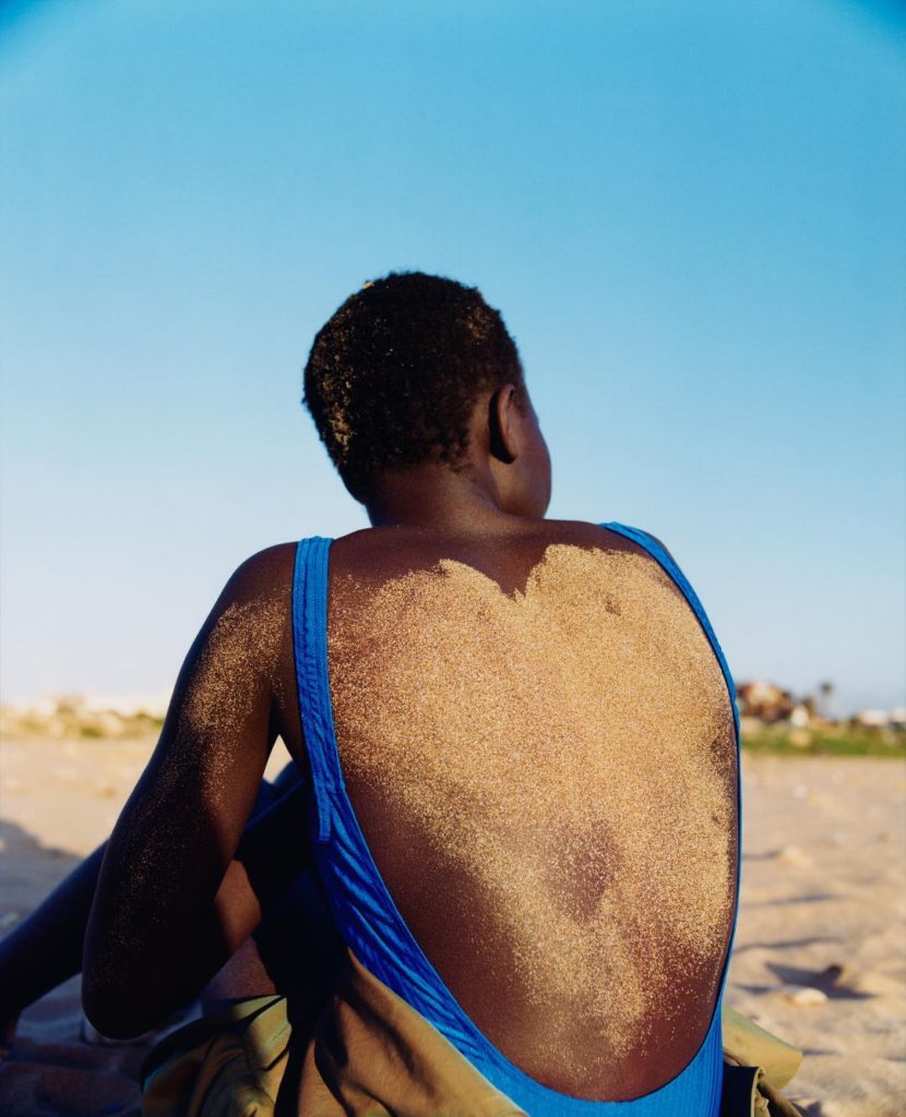 Tyler Mitchell photograph of the back of a women covered with sand wearing a blue one piece bathing suit sitting on a beach