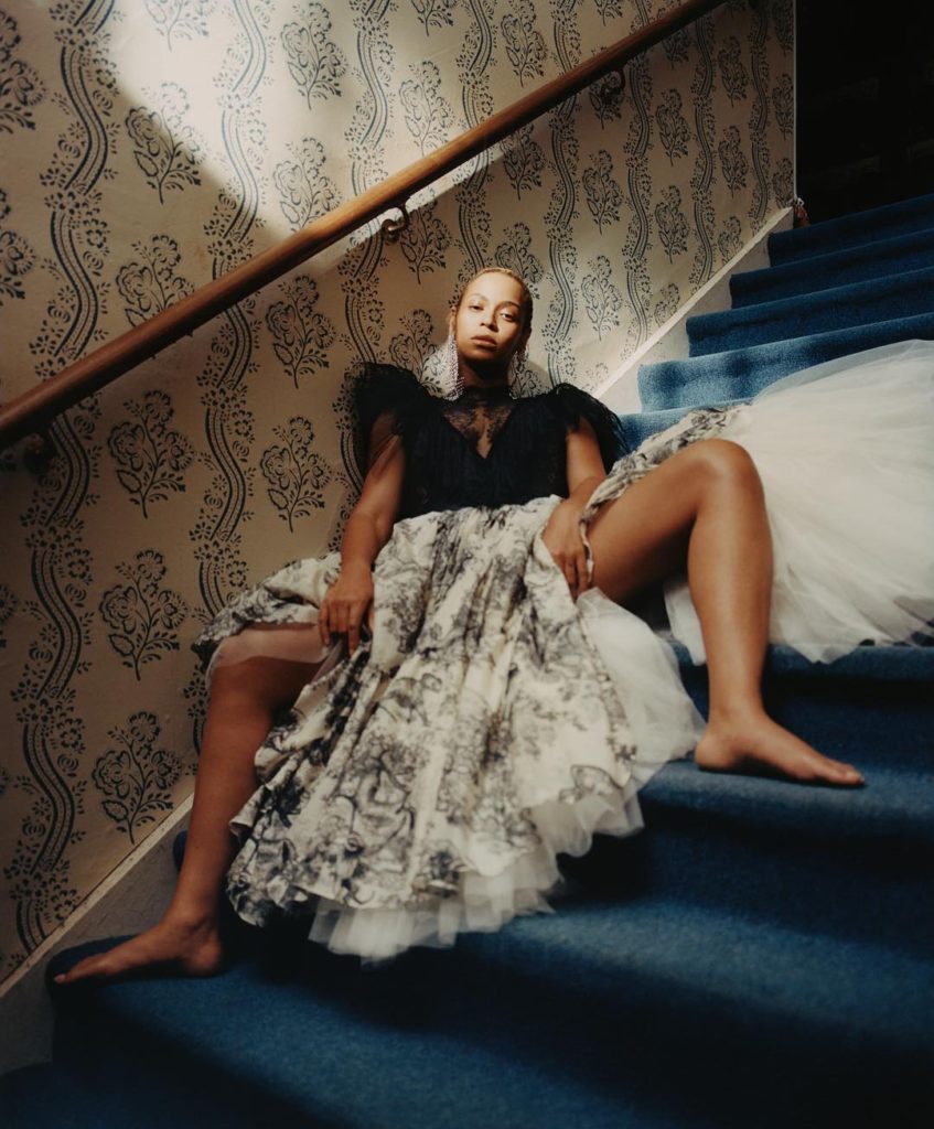 Tyler Mitchell Photograph of Beyonce Sitting on a Blue Carpeted Staircase with bare legs wide open wearing a full white and black printed hoop skirt
