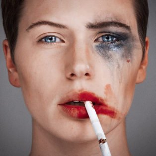 Tyler Shields photograph of a women with one side of her face with smeared make up and a cigarette dangling from her right side of her lip and the left side of her face all clean