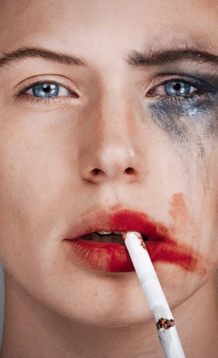 Tyler Shields photograph of a women with one side of her face with smeared make up and a cigarette dangling from her right side of her lip and the left side of her face all clean