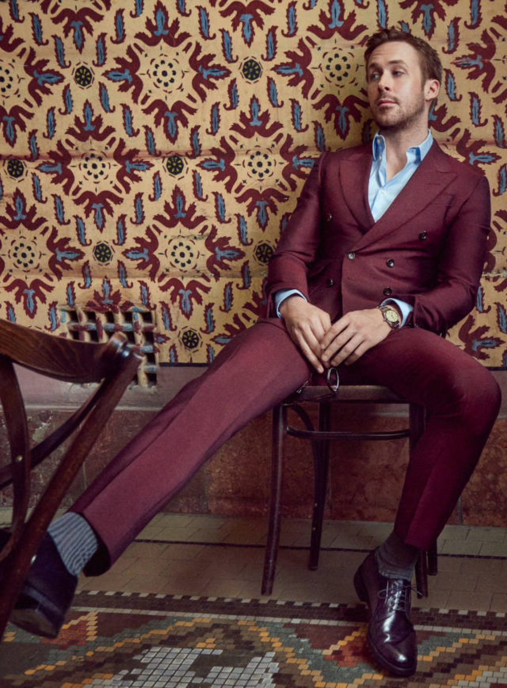 Actor Ryan Gosling in a Cranberry Double Breasted Suit sitting on a wooden cafe chair against a maroon and blue floral tapestry