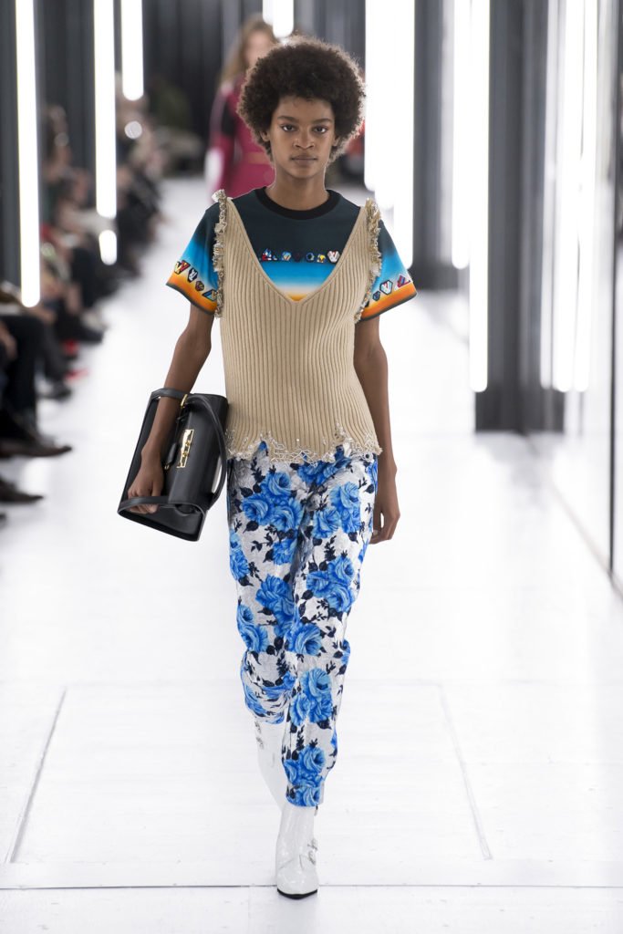 African American Runway Model Wearing Beige Vest, Black, Royal Blue and Orang Short-Sleeve Shirt, White Pants with Blue and Black Floral Pants and White Patent Leather Pointy Ankle Boots with Buckle