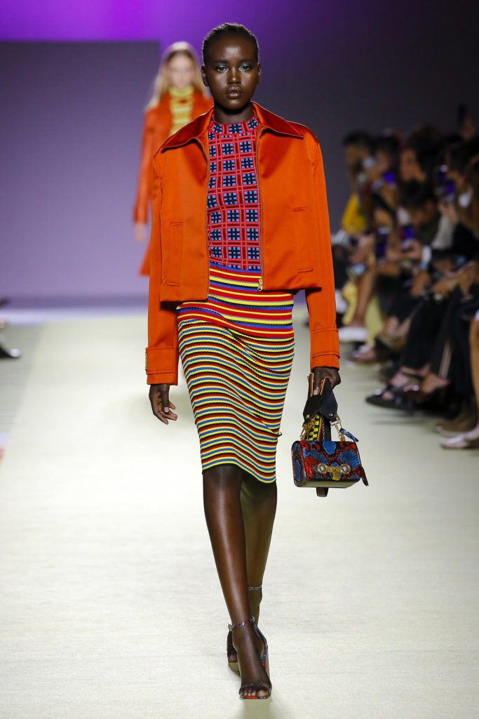 African American Female Runway Model wearing and orange jacket, red and blue block print top, red, yellow and blue pencil skirt carrying a mini tote
