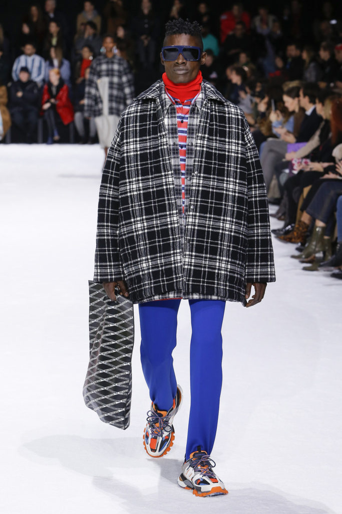 African American Male Runway Model Wearing Cool Blue Sunglasses, Red Turtleneck, red, blue and white striped shirt, black and white extra big shirt jacket, royal blue pants and orange and blue trainers