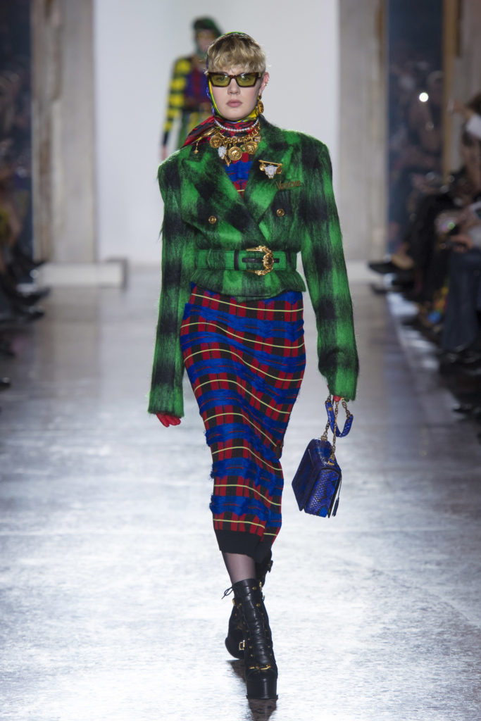Female Runway Model wearing green and black wool motorcycle jacket, blue, red, black and yellow dress, yellow and black sunglasses and gold necklace