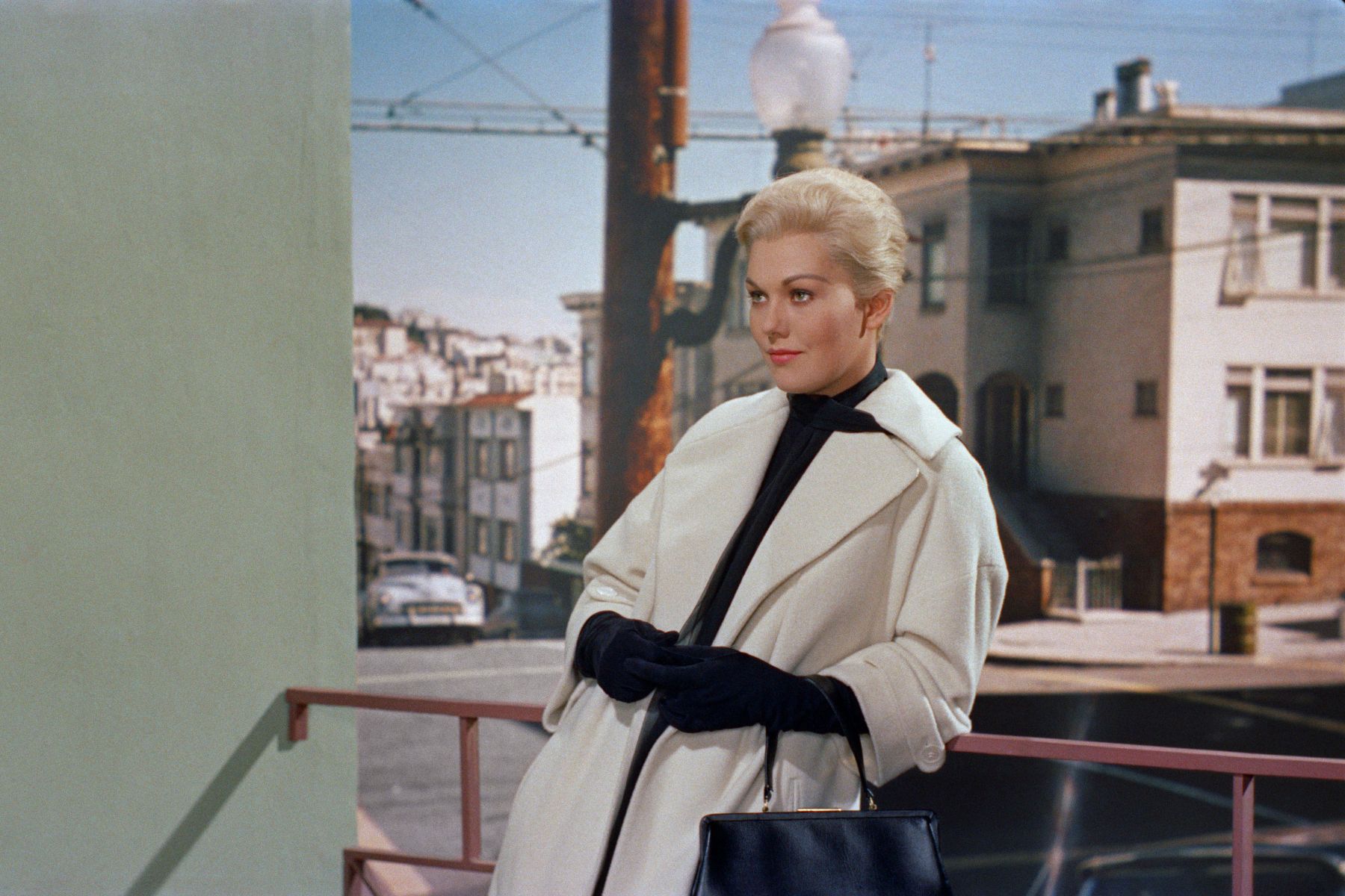 blonde, upperclass woman leans on a railing while wearing a white coat