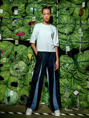 Eileen-Fisher and Public School Recycled Pants and Top on Model standing in front of a wall of stacked green laundry bags