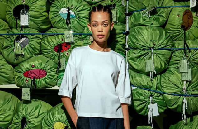 Eileen-Fisher and Public School Recycled Pants and Top on Model standing in front of a wall of stacked green laundry bags