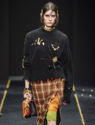 VERSACE Runway Fall 2019 Model wearing frayed black sweater with large V and other pins_plaid skirt with slit revealing an orange and lime green trimmed lace slip and lime green shoes