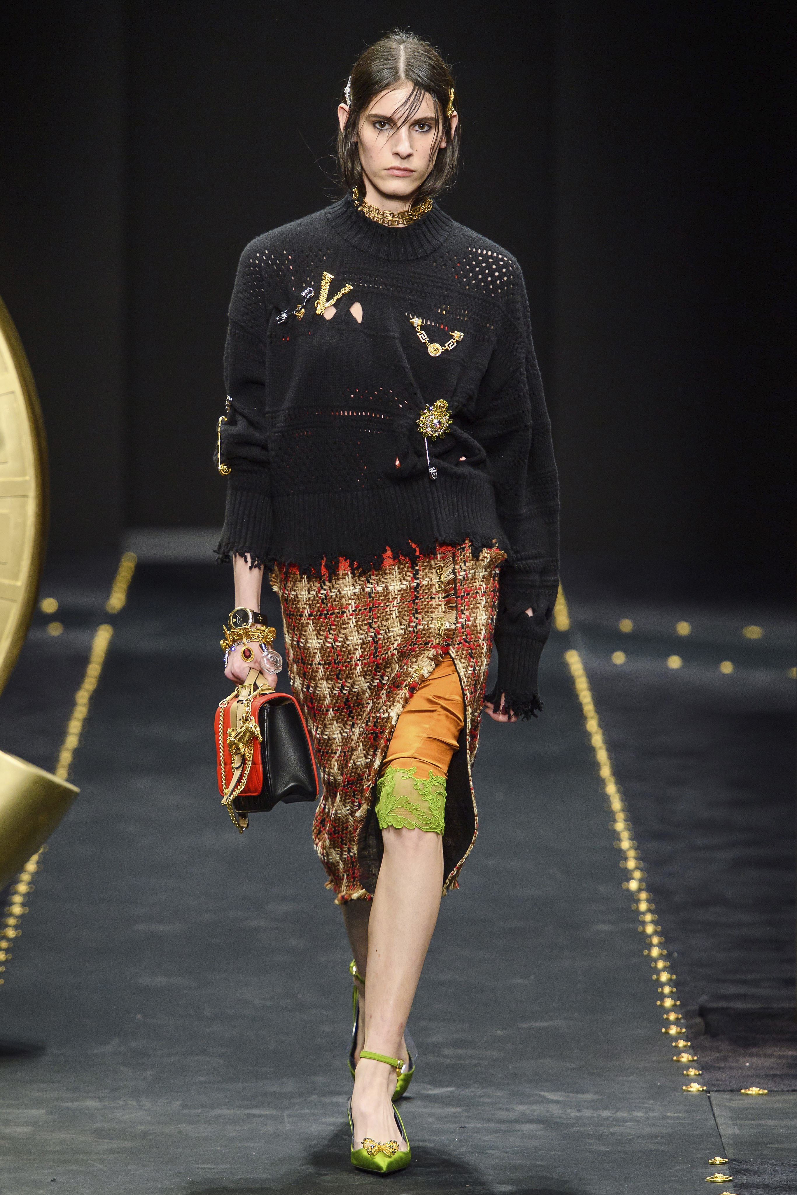 VERSACE Runway Fall 2019 Model wearing frayed black sweater with large V and other pins_plaid skirt with slit revealing an orange and lime green trimmed lace slip and lime green shoes