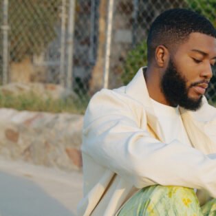 Khalid in white jacket sitting next to chain linked fence